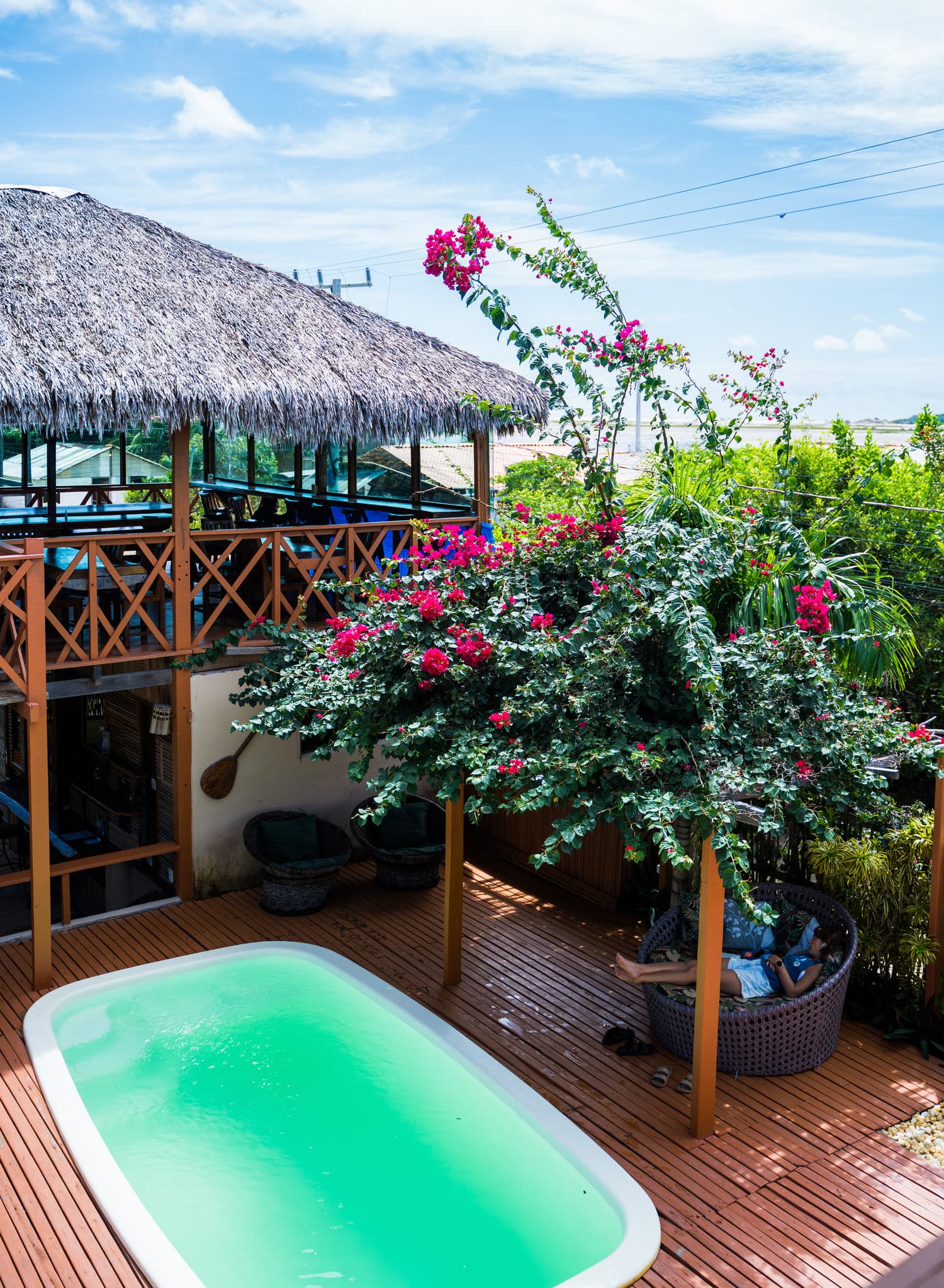 Piscina em deck de madeira sob buganvília, ao lado do quiosque de palha